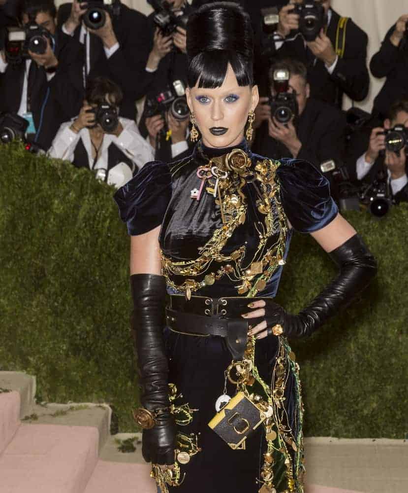 Katy Perry went dramatic wearing her gold embellished black gown and her dark hair was swept into a towering present day beehive with short bangs while wearing a bold make-up + eyebrow-free to complete the look at the 2016 MET Gala.