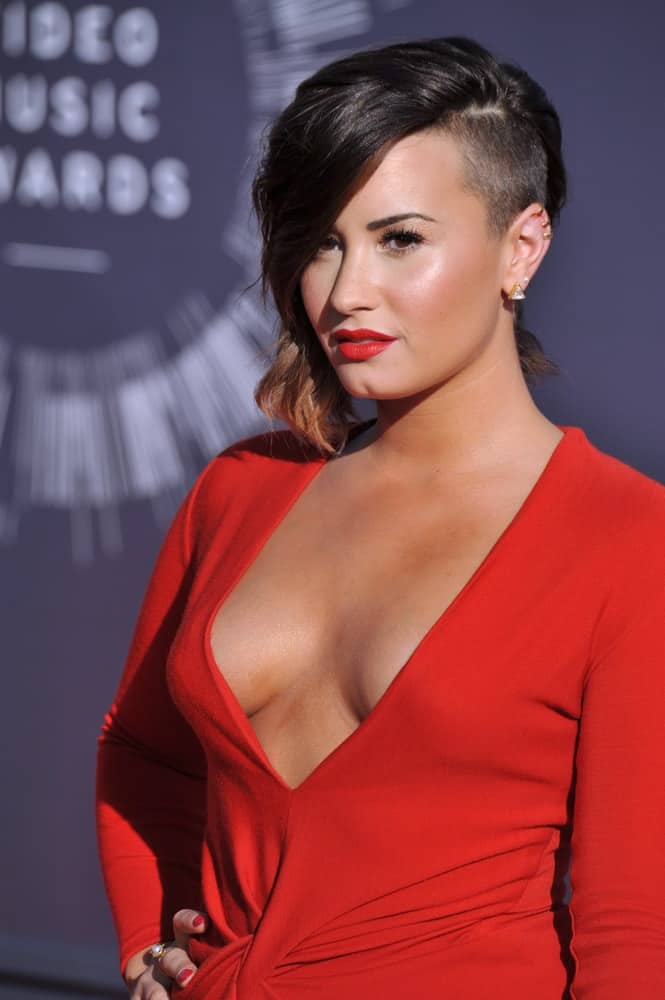 On August 24, 2014, Demi Lovato was quite lovely with her red dress, red lips and red nails to pair with her highlighted side-swept chin-length hairstyle with a shaved side of the head at the 2014 MTV Video Music Awards at the Forum in Los Angeles.