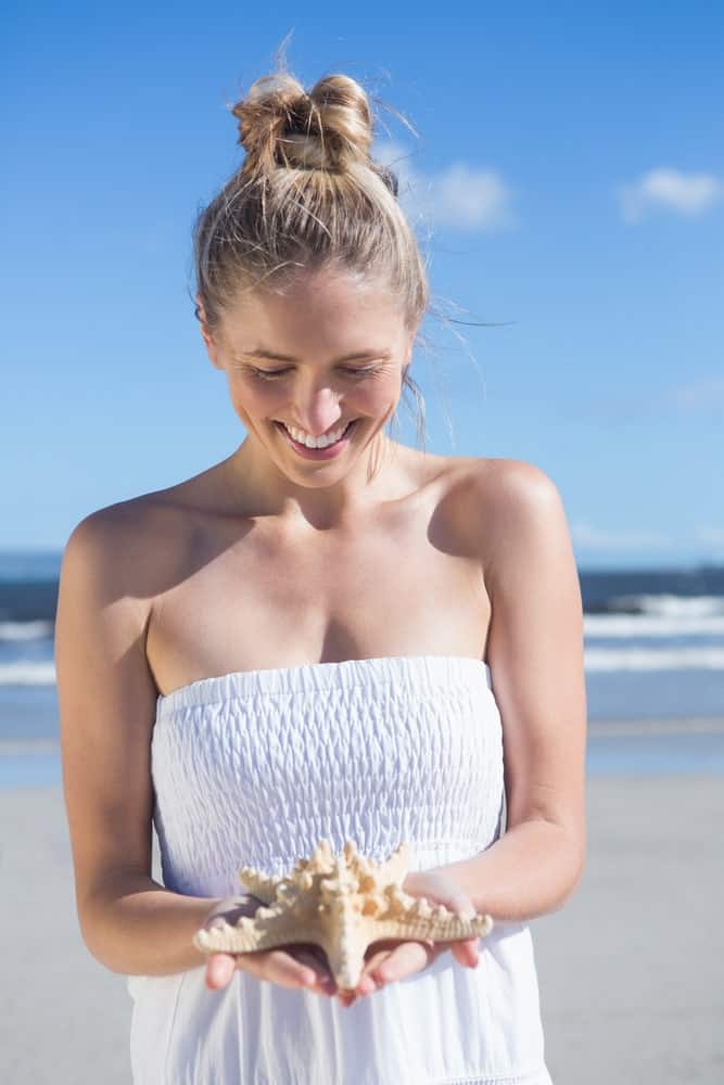 Woman in a white dress and a messy bun on the beach holding a starfish.